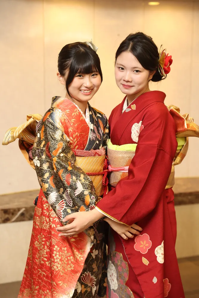 Two women wearing colorful kimono with gold obi sashes, smiling together indoors.