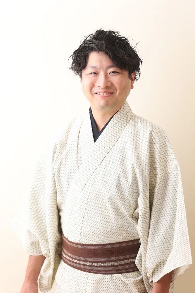 A man wearing a cream kimono with a brown obi, smiling.