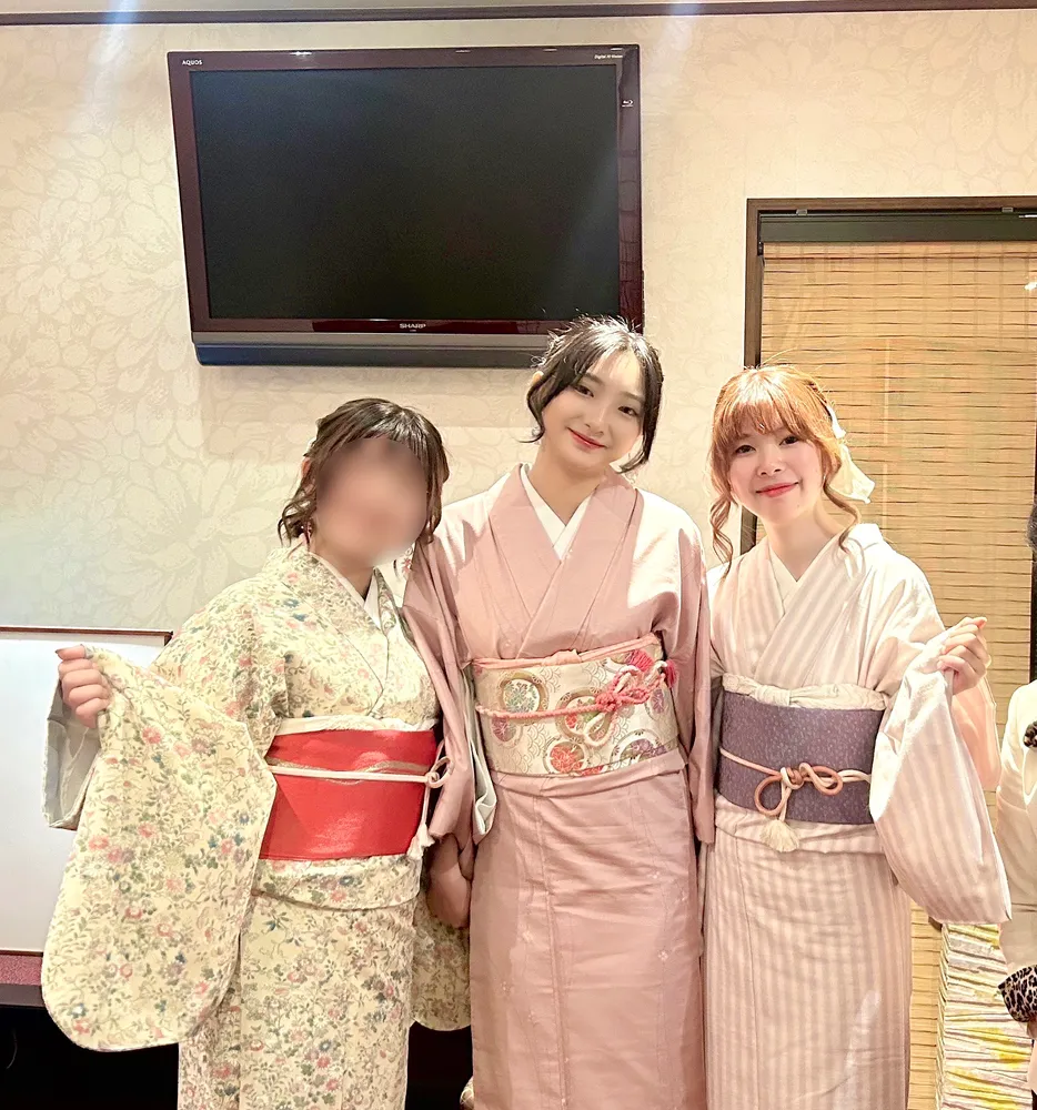 Three women in kimono of different patterns and colors (cream floral, plain pink, and lilac stripe), posing together indoors; one person's face is blurred for privacy.