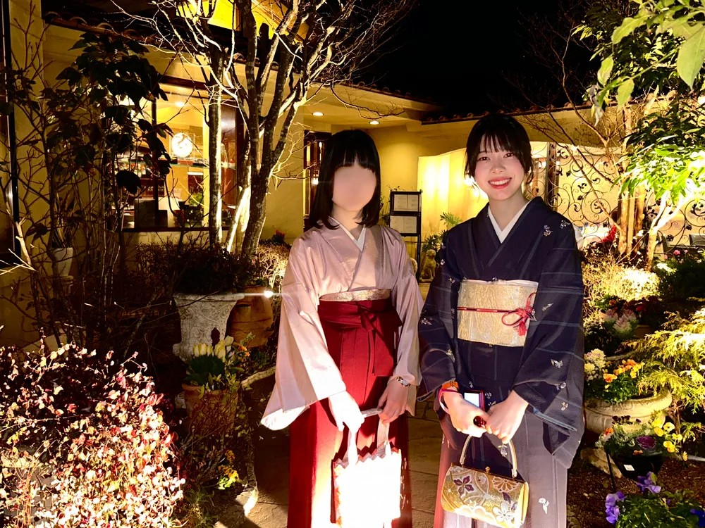 Two women in kimono outside a restaurant at night, one in pink with a red obi, one in navy with a gold obi; one person's face is blurred for privacy.