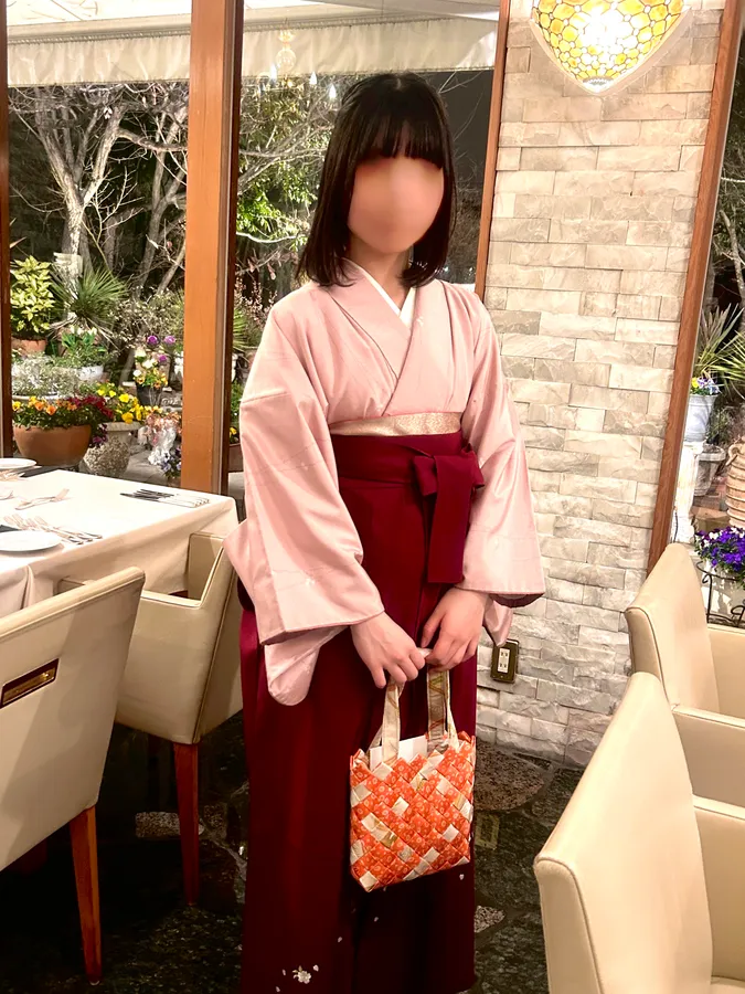 A woman in a pink kimono with a red obi holding a woven orange handbag, in a restaurant setting; her face is blurred for privacy.