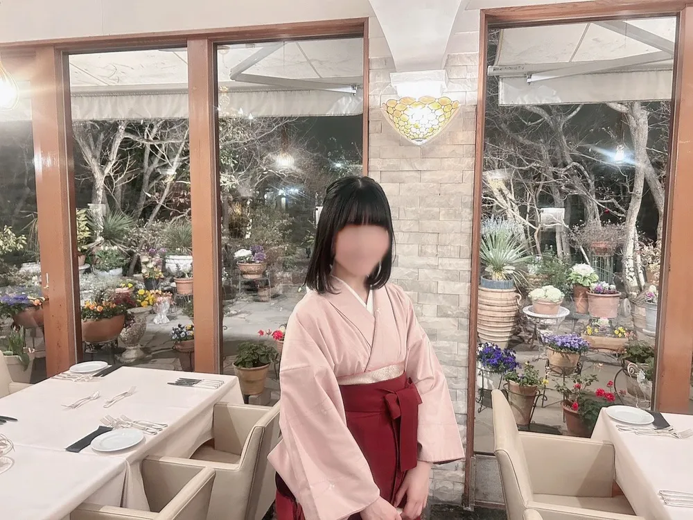 A woman in a pink kimono with a red obi standing in a garden at night; her face is blurred for privacy.