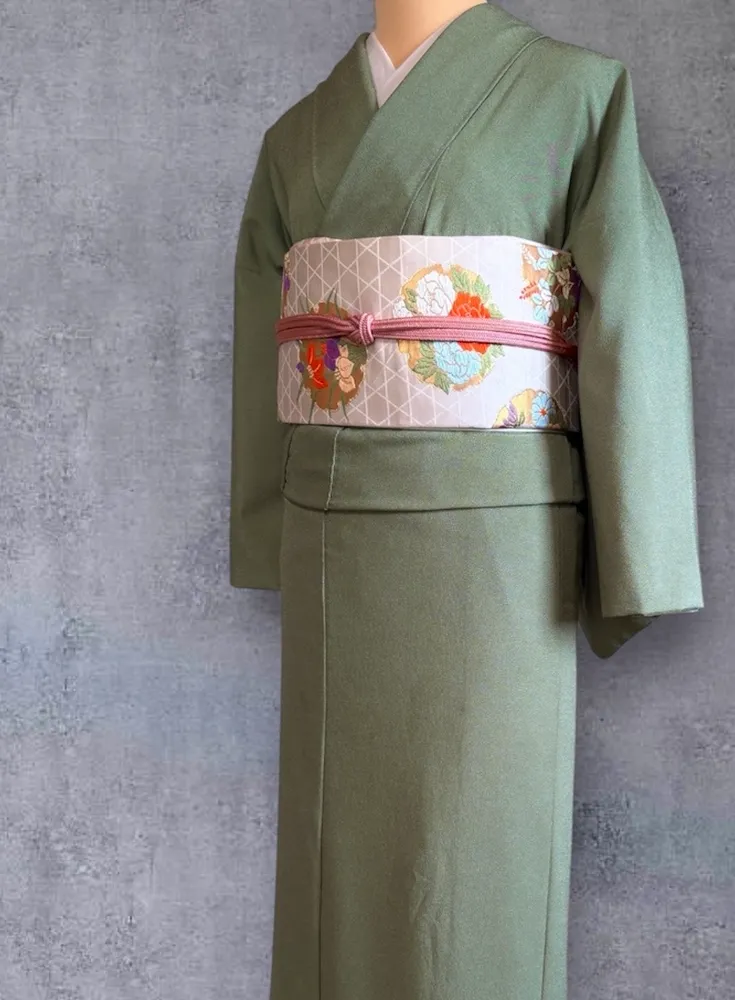 A sage green kimono on a mannequin, with a cream obi featuring floral embroidery.