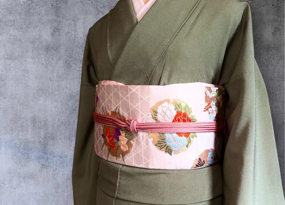Close-up of a cream obi with floral embroidery, tied with a dusty pink cord, over a sage green kimono.