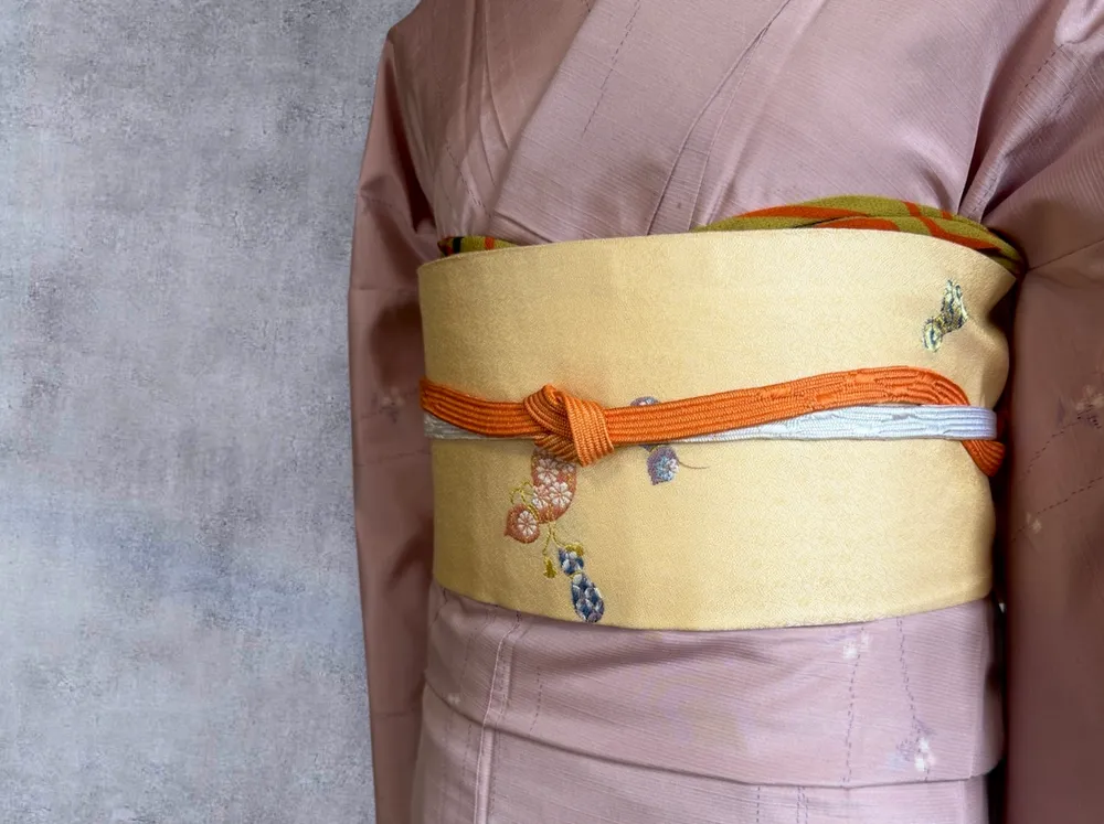 Close-up of a gold obi tied with an orange and white cord, over a dusty pink kimono.