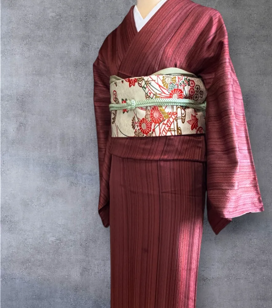 A burgundy striped kimono on a mannequin, tied with a cream obi featuring red and gold floral patterns.