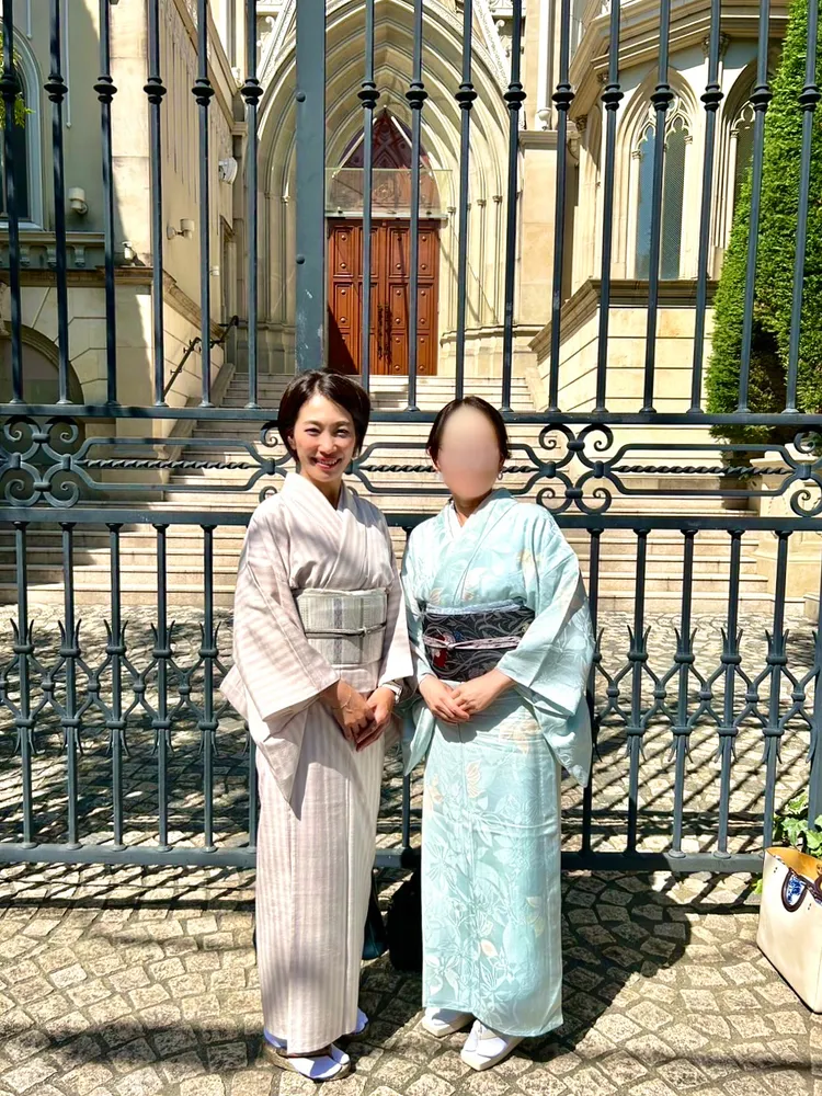 Two women in kimono standing in front of an ornate iron gate, one in beige stripes, one in pale blue with a floral pattern; one person's face is blurred for privacy.